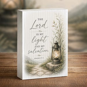 May include: A rectangular white wooden block sign featuring a printed illustration of a stone path, greenery, and a lit lantern. The text reads: The Lord is my light and my salvation. Psalm 27:1. The surface has a subtle vertical wood grain texture.