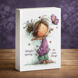 May include: A white wooden block features a whimsical illustration of a girl surrounded by butterflies. She wears a purple dress and green pants, with the text "Alright spring... do your thing!" The artwork has a watercolor style.