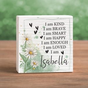 May include: A white wooden block with the words "I am KIND, BRAVE, SMART, HAPPY, ENOUGH, LOVED" and the name "Isabella" in script. The block is decorated with daisy flowers and green watercolor accents.