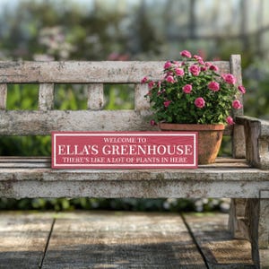 May include: A red wooden sign with white text that reads "Welcome to Ella's Greenhouse There's like a lot of plants in here." The sign is sitting on a white wooden bench with a potted plant in the background.