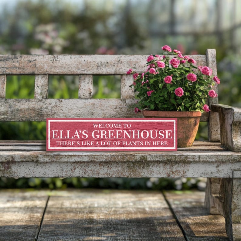 May include: A red wooden sign with white text that reads "Welcome to Ella's Greenhouse There's like a lot of plants in here." The sign is sitting on a white wooden bench with a potted plant in the background.