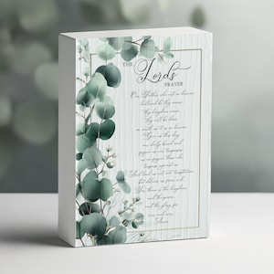 May include: A white wooden block featuring "The Lord's Prayer" in elegant script. The text is framed by a gold border and adorned with watercolor eucalyptus leaves and sprigs. The block is a decorative item.
