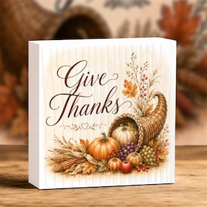 May include: A square wooden block sign featuring the text Give Thanks in elegant black script. The illustration displays a woven cornucopia overflowing with pumpkins, grapes, an apple, wheat stalks, and autumn leaves on a light wood background.