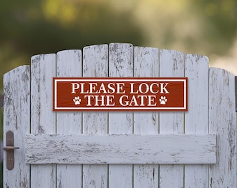 May include: A red wooden sign with white text that reads "PLEASE LOCK THE GATE" with paw prints on either side of the text. The sign is mounted on a white wooden fence.