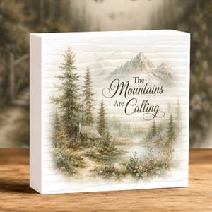 May include: A rectangular white wooden block sign featuring a printed landscape illustration of a cabin, pine trees, a lake, and mountains. The text "The Mountains Are Calling" is printed in black script and serif font on the right side.