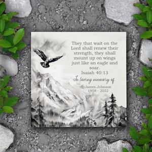 May include: A square memorial plaque with a black and white illustration of an eagle soaring above a mountain range. Text includes a Bible verse from Isaiah 40:13 and the inscription "In loving memory of James Johnson 1958-2022."