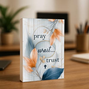May include: A rectangular wooden block with the words "pray wait... trust" in a script font. The design features orange and blue botanical elements, gold circles, and a small cross. The background has a light blue and white wood grain pattern.