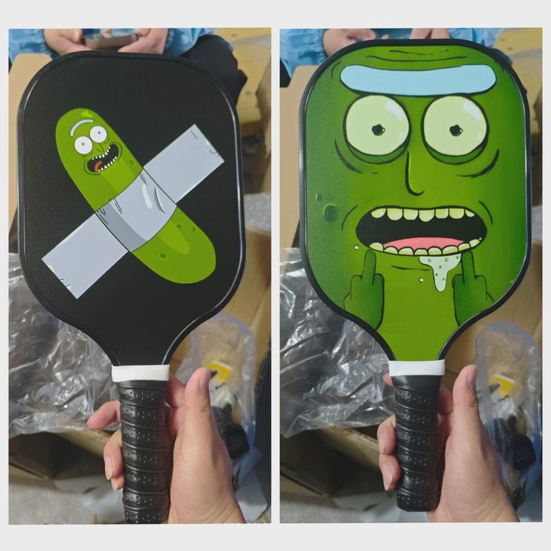 K&ouml;nnte beinhalten: Zwei Pickleball-Schl&auml;ger mit Cartoon-Charakter-Designs. Ein Schl&auml;ger zeigt eine Gurke mit Klebeband, der andere ein gr&uuml;nes Gesicht mit erhobenen Mittelfingern. Beide haben schwarze Griffe mit wei&szlig;en Akzenten.