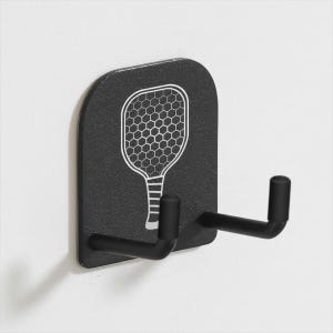 May include: Black wall-mounted hook with a white pickleball paddle design. The hook has two arms extending from a rectangular base. The paddle graphic features a honeycomb pattern. The hook is mounted on a white wall.
