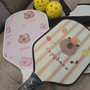 K&ouml;nnte beinhalten: Zwei Pickleball-Schl&auml;ger mit Capybara-Designs, einer rosa mit einem B&auml;ren und dem Text "Cadybara", der andere mit Pfirsichstreifen und einem B&auml;ren, dem Text "capybara" und zwei gelben Pickleballs. Die Schl&auml;ger haben schwarze Griffe.