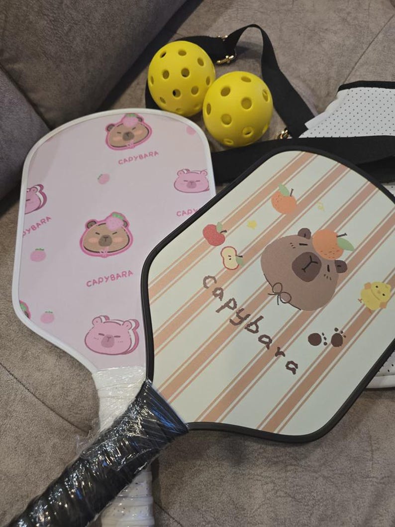 K&ouml;nnte beinhalten: Zwei Pickleball-Schl&auml;ger mit Capybara-Designs, einer rosa mit einem B&auml;ren und dem Text "Cadybara", der andere mit Pfirsichstreifen und einem B&auml;ren, dem Text "capybara" und zwei gelben Pickleballs. Die Schl&auml;ger haben schwarze Griffe.