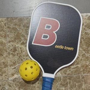 K&ouml;nnte beinhalten: Ein schwarzer Pickleball-Schl&auml;ger mit wei&szlig;em Rand und einem gro&szlig;en roten und wei&szlig;en Buchstaben "B" auf der Vorderseite. Der Text "eetle town" ist in Gold gedruckt. Ein gelber, perforierter Pickleball liegt neben dem Schl&auml;ger. Der Griff ist blau.