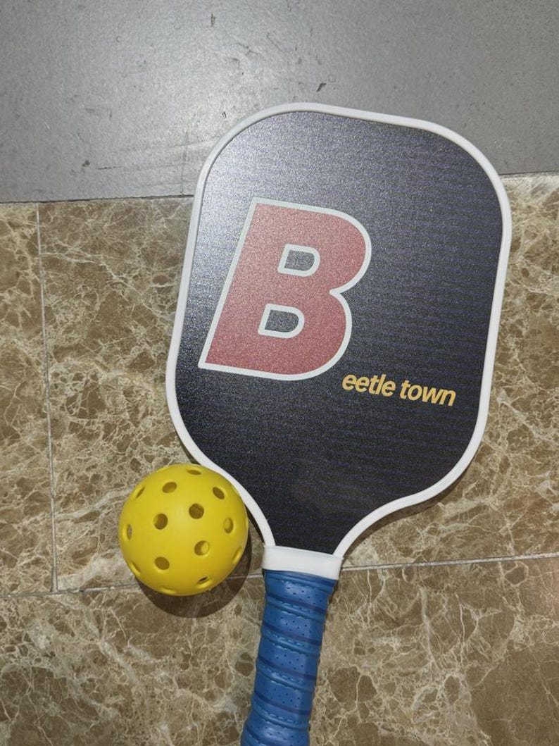 K&ouml;nnte beinhalten: Ein schwarzer Pickleball-Schl&auml;ger mit wei&szlig;em Rand und einem gro&szlig;en roten und wei&szlig;en Buchstaben "B" auf der Vorderseite. Der Text "eetle town" ist in Gold gedruckt. Ein gelber, perforierter Pickleball liegt neben dem Schl&auml;ger. Der Griff ist blau.