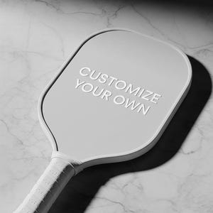 K&ouml;nnte beinhalten: Ein hellgrauer Pickleball-Schl&auml;ger mit dem wei&szlig;en Text "CUSTOMIZE YOUR OWN". Der Schl&auml;ger hat einen strukturierten wei&szlig;en Griff und einen abgerundeten Kopf. Der Hintergrund ist ein Marmormuster.