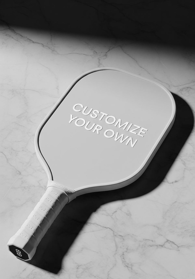 K&ouml;nnte beinhalten: Ein hellgrauer Pickleball-Schl&auml;ger mit dem wei&szlig;en Text "CUSTOMIZE YOUR OWN". Der Schl&auml;ger hat einen strukturierten wei&szlig;en Griff und einen abgerundeten Kopf. Der Hintergrund ist ein Marmormuster.
