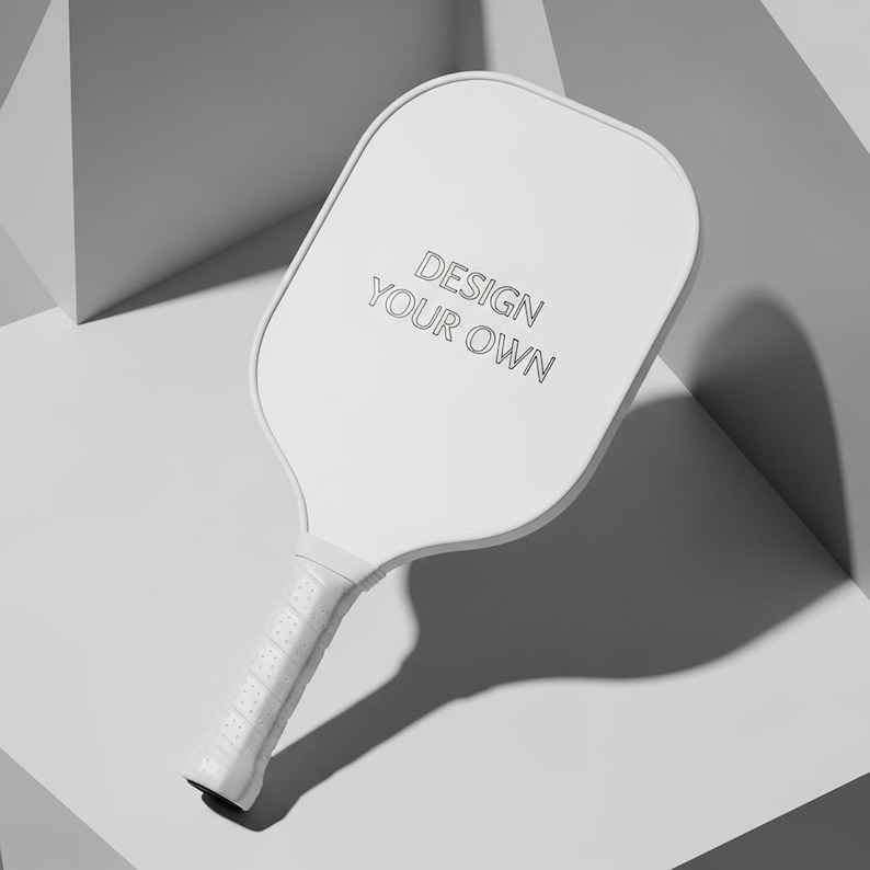 K&ouml;nnte beinhalten: Ein wei&szlig;er Pickleball-Schl&auml;ger mit dem Text "DESIGN YOUR OWN" auf der Schl&auml;gerfl&auml;che. Der Griff hat eine strukturierte Oberfl&auml;che. Der Schl&auml;ger befindet sich vor einem wei&szlig;en und grauen geometrischen Hintergrund.