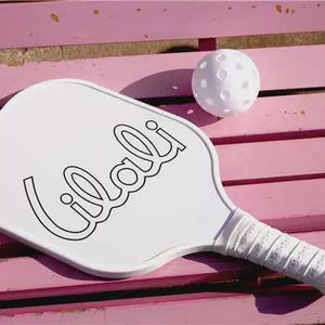 K&ouml;nnte beinhalten: Ein wei&szlig;er Pickleball-Schl&auml;ger mit dem Wort "Lilali" in schwarzer Schrift und einem wei&szlig;en, perforierten Griff. Ein wei&szlig;er Pickleball liegt neben dem Schl&auml;ger. Die Artikel befinden sich auf einer rosa Holzoberfl&auml;che.