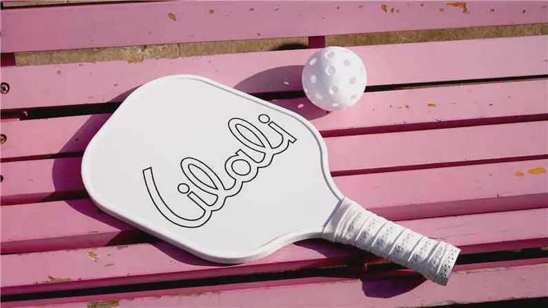 K&ouml;nnte beinhalten: Ein wei&szlig;er Pickleball-Schl&auml;ger mit dem Wort "Lilali" in schwarzer Schrift und einem wei&szlig;en, perforierten Griff. Ein wei&szlig;er Pickleball liegt neben dem Schl&auml;ger. Die Artikel befinden sich auf einer rosa Holzoberfl&auml;che.