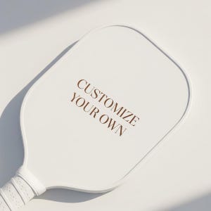 K&ouml;nnte beinhalten: Ein wei&szlig;er Pickleball-Schl&auml;ger mit strukturiertem Griff und dem Text "CUSTOMIZE YOUR OWN" in Braun. Der Schl&auml;ger befindet sich vor einem wei&szlig;en Hintergrund mit weichen Schatten, was eine saubere, moderne &Auml;sthetik suggeriert.