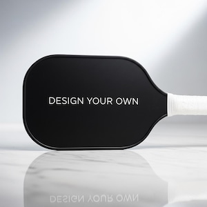 K&ouml;nnte beinhalten: Ein schwarzer Pickleball-Schl&auml;ger mit dem wei&szlig;en Text "DESIGN YOUR OWN". Der Griff ist mit wei&szlig;em Griffband umwickelt. Der Schl&auml;ger liegt auf einer wei&szlig;en und grauen Oberfl&auml;che.