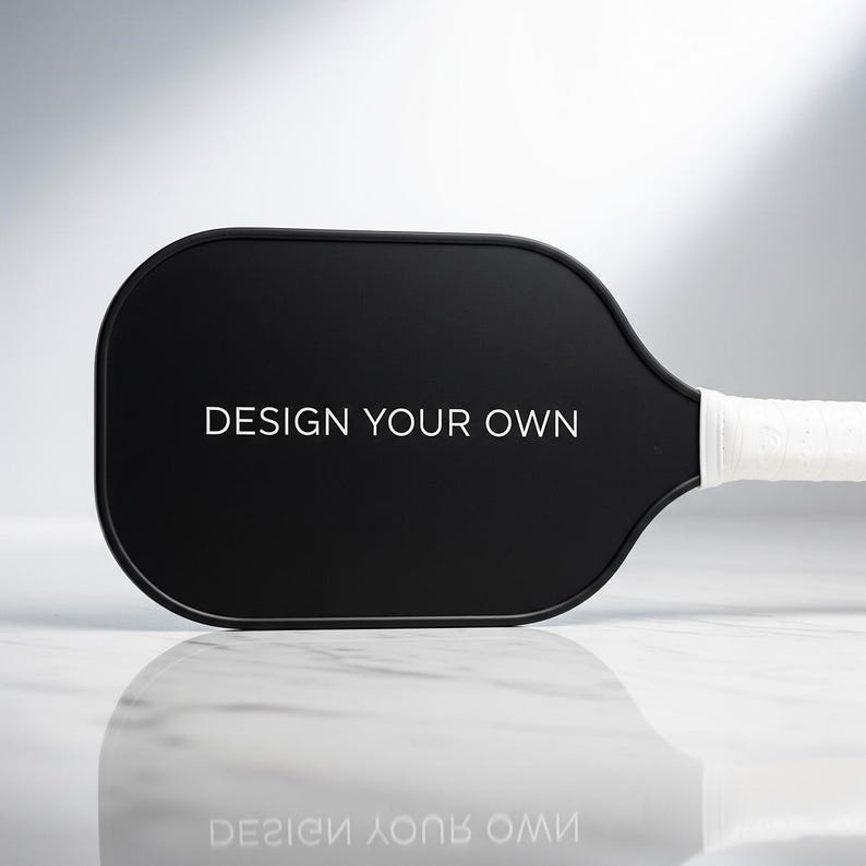 K&ouml;nnte beinhalten: Ein schwarzer Pickleball-Schl&auml;ger mit dem wei&szlig;en Text "DESIGN YOUR OWN". Der Griff ist mit wei&szlig;em Griffband umwickelt. Der Schl&auml;ger liegt auf einer wei&szlig;en und grauen Oberfl&auml;che.