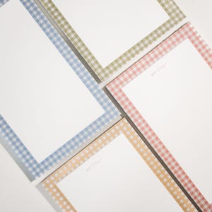 May include: A collection of notepads with white pages and gingham patterned borders in blue, green, pink, and yellow. The word "notes" is written in cursive in the upper left corner of each notepad.