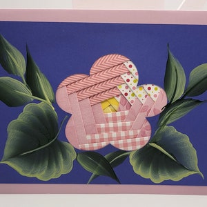 May include: A handmade greeting card featuring a pink flower with a woven paper design, set against a deep blue background. The flower is surrounded by green painted leaves, all framed by a pink border.