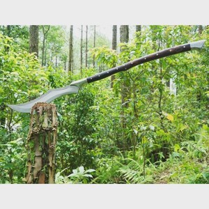 May include: A large, silver-bladed weapon with a curved edge, set on a wooden handle with a dark brown finish. The weapon is displayed outdoors, resting on a tree stump, with a lush green forest in the background.