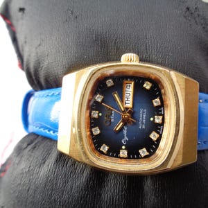 Peut inclure: Une montre-bracelet vintage dorée avec un cadran carré et un bracelet en cuir bleu. Le cadran est bleu foncé avec des marqueurs en forme de diamant et des aiguilles dorées. Le jour est affiché comme "THU 4". La montre est étiquetée "Crystal" et "Star".