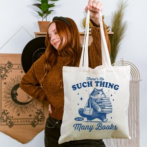 Peut inclure: Sac fourre-tout cr&egrave;me avec une illustration bleue d'un chat portant des livres. Le texte sur le sac se lit &laquo; There's No Such Thing As Too Many Books &raquo;. Le sac a de longues anses et est tenu.