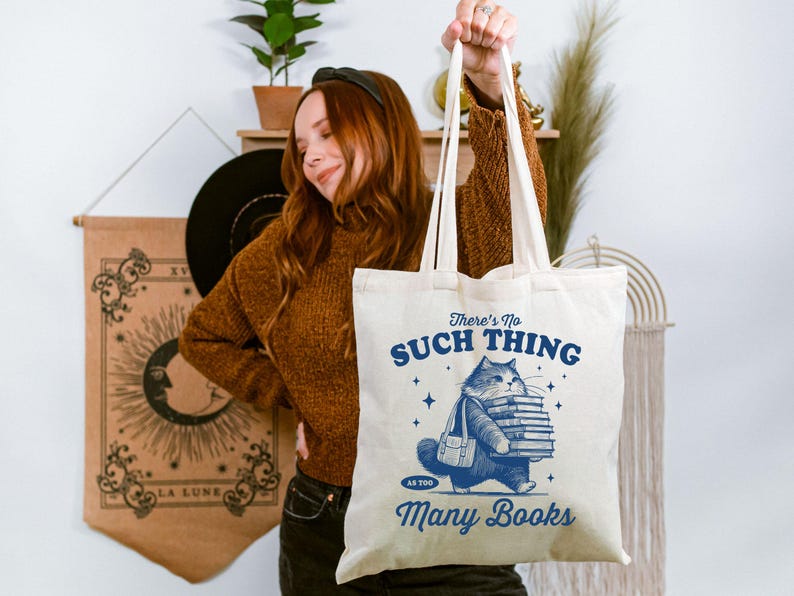 Peut inclure: Sac fourre-tout cr&egrave;me avec une illustration bleue d'un chat portant des livres. Le texte sur le sac se lit &laquo; There's No Such Thing As Too Many Books &raquo;. Le sac a de longues anses et est tenu.