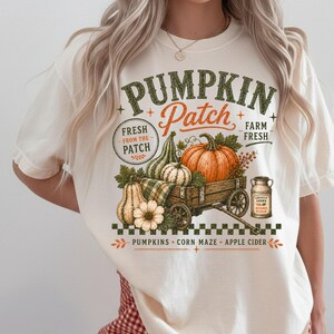 May include: An off-white t-shirt featuring a graphic of a wooden wagon filled with pumpkins and gourds. The text reads PUMPKIN Patch, FRESH FROM THE PATCH, FARM FRESH, LOCALLY GROWN, AUTUMN PICKED, PUMPKINS, CORN MAZE, and APPLE CIDER with green accents.