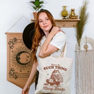 Peut inclure: Sac fourre-tout cr&egrave;me avec un chat portant des livres et le texte &laquo;&nbsp;There's No Such Thing As Too Many Books&nbsp;&raquo;. Le sac est tenu par une personne portant un t-shirt blanc.
