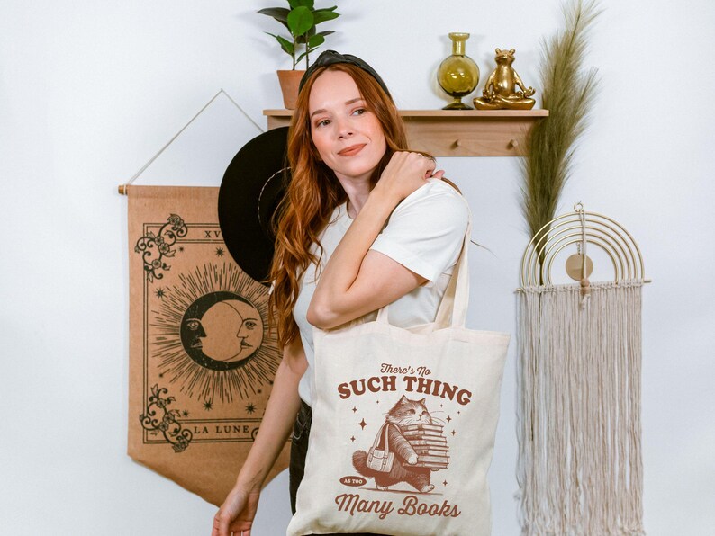 Peut inclure: Sac fourre-tout cr&egrave;me avec un chat portant des livres et le texte &laquo;&nbsp;There's No Such Thing As Too Many Books&nbsp;&raquo;. Le sac est tenu par une personne portant un t-shirt blanc.