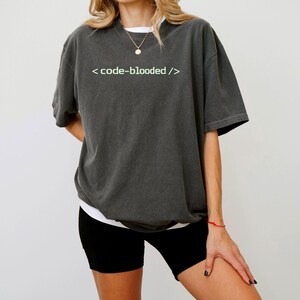 K&ouml;nnte beinhalten: Dunkelgraues T-Shirt mit dem wei&szlig;en Text "< code-blooded />". Das Shirt hat kurze &Auml;rmel und eine lockere Passform. Das Model tr&auml;gt schwarze Shorts und ein wei&szlig;es Unterhemd.