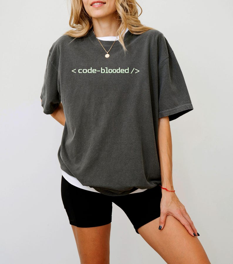 K&ouml;nnte beinhalten: Dunkelgraues T-Shirt mit dem wei&szlig;en Text "< code-blooded />". Das Shirt hat kurze &Auml;rmel und eine lockere Passform. Das Model tr&auml;gt schwarze Shorts und ein wei&szlig;es Unterhemd.
