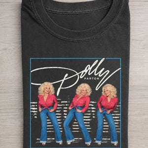 May include: A charcoal gray crew neck t-shirt featuring a graphic print of Dolly Parton in a pink button-down shirt and blue jeans. The design includes the white cursive text Dolly Parton enclosed within a thin blue rectangular border.