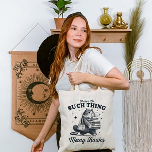 Peut inclure: Sac fourre-tout cr&egrave;me avec un chat portant des livres et le texte "There's No Such Thing as Too Many Books". Le chat porte une sacoche. Le sac est tenu par une personne.