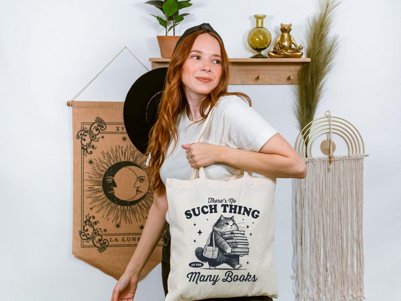 Peut inclure: Sac fourre-tout cr&egrave;me avec un chat portant des livres et le texte "There's No Such Thing as Too Many Books". Le chat porte une sacoche. Le sac est tenu par une personne.