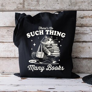 Peut inclure: Sac fourre-tout noir avec une illustration de chat portant des livres. Le texte sur le sac dit "There's No Such Thing As Too Many Books" en blanc. Le chat et les livres sont en noir et blanc.