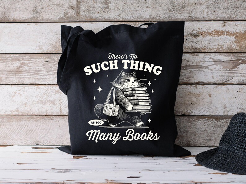 Peut inclure: Sac fourre-tout noir avec une illustration de chat portant des livres. Le texte sur le sac dit "There's No Such Thing As Too Many Books" en blanc. Le chat et les livres sont en noir et blanc.
