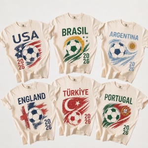 Peut inclure: Six t-shirts crème avec des motifs de ballon de football et les noms de pays : USA, Brasil, Argentina, England, Türkiye et Portugal. Chaque t-shirt affiche l'année 2026 sous le ballon.