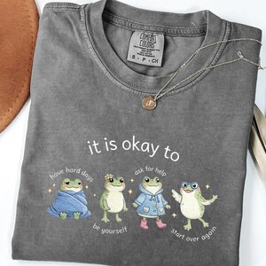 Op de afbeelding: Een grijs Comfort Colors T-shirt met de zin "it is okay to" en kikkerillustraties. Het shirt heeft de zinnen "have hard days", "be yourself", "ask for help" en "start over again". Een gouden ketting is over het shirt gedrapeerd.