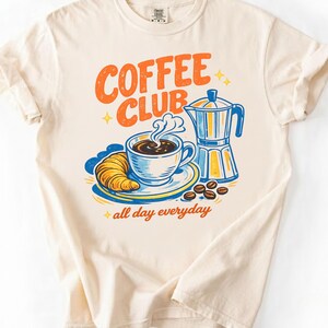 May include: A cream-coloured t-shirt with a colourful graphic. The graphic shows the words "COFFEE CLUB" in orange, a cup of coffee, a croissant, and a coffee maker. The phrase "all day everyday" is also included.