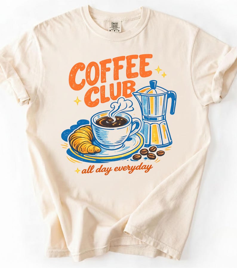May include: A cream-coloured t-shirt with a colourful graphic. The graphic shows the words "COFFEE CLUB" in orange, a cup of coffee, a croissant, and a coffee maker. The phrase "all day everyday" is also included.