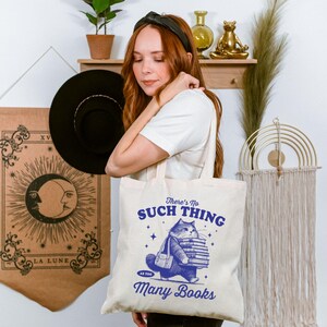 Peut inclure: Sac fourre-tout cr&egrave;me avec une illustration bleue d'un chat portant des livres et le texte "There's No Such Thing as Too Many Books". Une personne tient le sac. Un chapeau noir et une tapisserie avec un motif de lune sont en arri&egrave;re-plan.