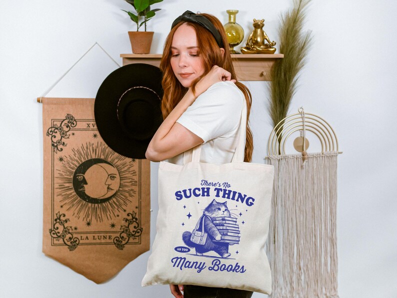 Peut inclure: Sac fourre-tout cr&egrave;me avec une illustration bleue d'un chat portant des livres et le texte "There's No Such Thing as Too Many Books". Une personne tient le sac. Un chapeau noir et une tapisserie avec un motif de lune sont en arri&egrave;re-plan.