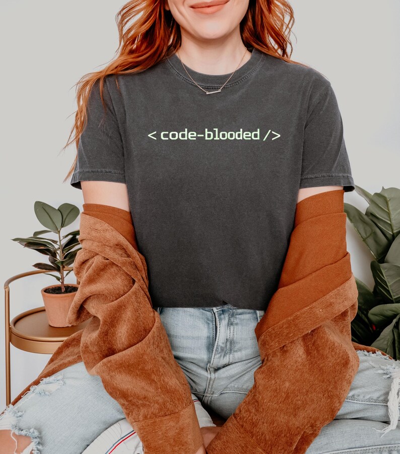 K&ouml;nnte beinhalten: Dunkelgraues T-Shirt mit dem Text "< code-blooded />" in Hellgr&uuml;n. Das Shirt wird mit einer braunen Jacke und blauen Jeans getragen. Das Thema bezieht sich auf das Programmieren und die Informatik.