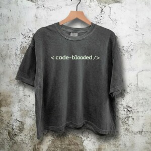 K&ouml;nnte beinhalten: Dunkelgraues T-Shirt mit dem wei&szlig;en Text "< code-blooded />". Das Shirt h&auml;ngt an einem Holzb&uuml;gel vor einer strukturierten, cremefarbenen Wand. Das T-Shirt hat kurze &Auml;rmel und einen Rundhalsausschnitt.