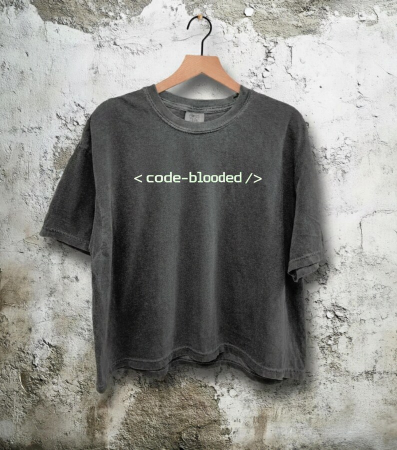 K&ouml;nnte beinhalten: Dunkelgraues T-Shirt mit dem wei&szlig;en Text "< code-blooded />". Das Shirt h&auml;ngt an einem Holzb&uuml;gel vor einer strukturierten, cremefarbenen Wand. Das T-Shirt hat kurze &Auml;rmel und einen Rundhalsausschnitt.